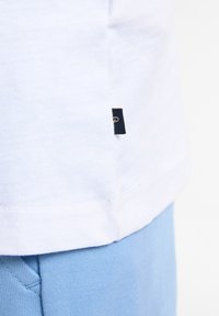Camiseta blanca de algodón con una etiqueta en la costura lateral y un dobladillo corto y recto, combinada con una tela azul clara que presenta una textura suave.