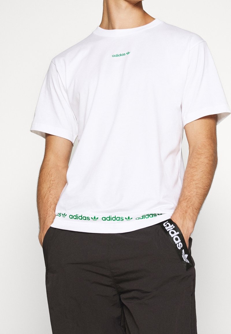 Vit bomull t-shirt med grön "adidas" logotyp på bröstet och en grön accentrand vid nederkanten. Rund hals, korta ärmar, avslappnad passform.