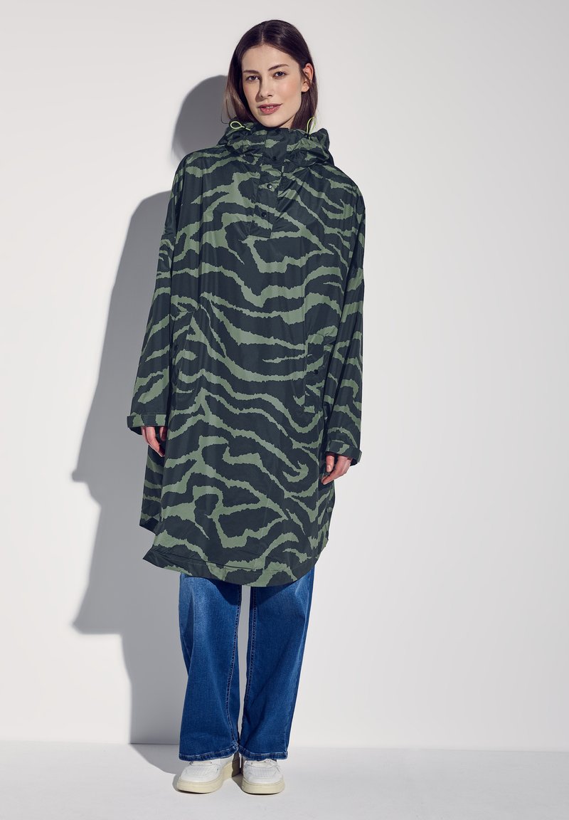 Lång huvad regnjacka i grönt med ett svart tigerrandigt mönster, tillverkad av ett lätt material, ihop med lösa blå jeans och vita sneakers.