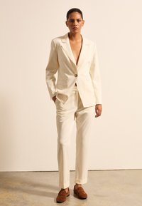 Helleckiger, taillierter Anzug mit tiefem V-Ausschnitt-Blazer und schmal zulaufenden Hosen. Verfügt über einen einzelnen Knopf, elegante Linien und einen subtilen Glanz. Braune Wildlederschuhe.