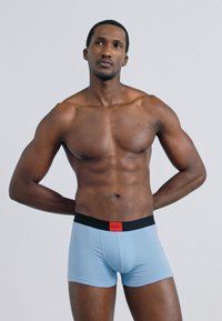 Ανοιχτό μπλε μπόξερ σλιπ με μαύρη μέση που φέρει κόκκινο λογότυπο. Λείο υλικό και εφαρμοστή γραμμή. Μοντέλο σε ουδέτερη στάση.