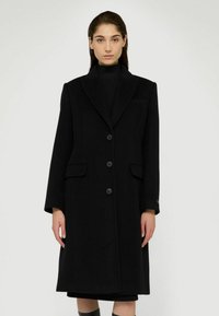 Černý vlněný kabát s dlouhým, rovnými liniemi, oversized límcem, třemi knoflíky a bočními kapsami, který se vyznačuje hladkou texturou.