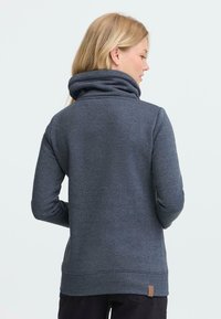 Mørk grå hettegenser med høy krage, tettsittende ermer og teksturert stoff. Har et lite brunt lærmerke på nedre del av ryggen.