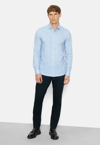 Helles blaues Hemd mit langen Ärmeln, subtiler Textur, kombiniert mit dunkelblauen Hosen und schwarzen Schuhen. Das Model steht vor einem einfarbigen Hintergrund.