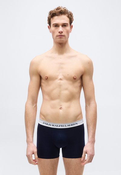 Νεαρός άνδρας όρθιος, χωρίς πουκάμισο, φορώντας σκούρα μποξεράκια με λευκή μέση που φέρει τη λέξη "Polo Ralph Lauren" σε απλό φόντο.