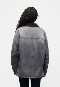 Άτομο με μακριά κατσαρά μαλλιά που φοράει μια ξεθωριασμένη μαύρη τζιν μπουφάν με πιο σκούρο γιακά, στέκεται με την πλάτη προς τα εμπρός μπροστά σε ένα απλό φωτεινό φόντο.