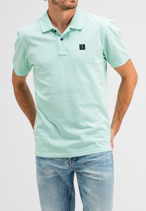 Polo vert menthe avec un plastron à deux boutons, manches courtes et un patch logo noir. Porté avec un jean bleu clair. Texture lisse.