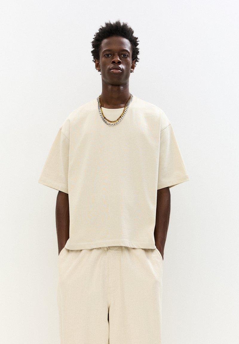 Tricou oversized bej cu mâneci scurte pliate, asortat cu pantaloni bej. Textură de țesătură netedă și o croială relaxată.