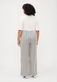 Eine Person in Übergröße trägt ein weißes Oberteil und gestreifte, weit geschnittene Hosen und wird von hinten gesehen, was einen entspannten, stilvollen Look zeigt.