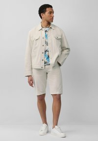 Lichtbeige spijkerjas over een blauw-witte tropische printshirt, gecombineerd met lichtbeige shorts en witte sneakers. Eenvoudig ontwerp, casual pasvorm.