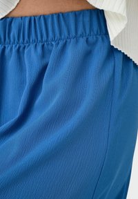 Abbigliamento blu e strutturato con una vita elastica. Il tessuto morbido cade delicatamente, creando una vestibilità rilassata. Nessun motivo o hardware visibile.