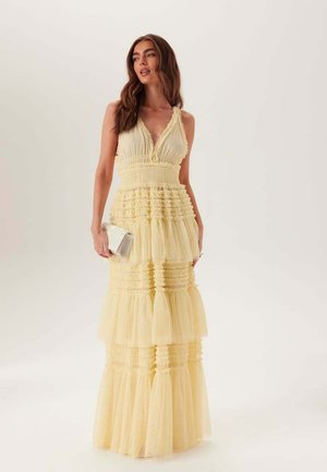 Kvinna med långt brunt hår som bär en ärmlös gul maxi-klänning med volanger, håller en liten vit kuvertväska, stående mot en enfärgad bakgrund.