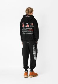 Persona con cabello rizado vistiendo una sudadera negra con capucha y pantalones con gráficos y texto en rojo, blanco y negro, de pie contra un fondo blanco.