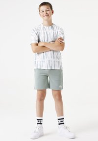 Un joven lleva una camiseta de manga corta de teñido anudado en blanco y azul, combinada con pantalones cortos verdes y zapatillas blancas. Medias a rayas en blanco y negro.