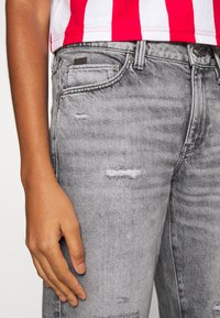 Vaqueros grises rasgados con parches desgastados, diseño clásico de cinco bolsillos y una sutil textura de denim, combinados con una blusa a rayas rojas y blancas.
