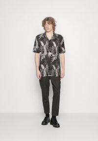 Musta lyhythihainen paita, jossa on valkoinen lehtikuvio, yhdistettynä tummiin housuihin ja mustiin kenkiin. Sisältää klassisen kauluksen ja rentoa istuvuutta.