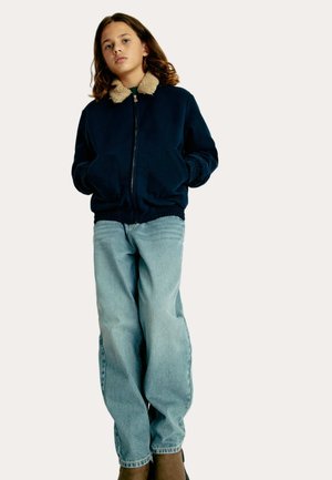 Marineblauwe ritsjack met een beige nepbont kraag, voorzien van twee voorzakken, gedragen over lichtblauwe jeans en bruine laarzen.