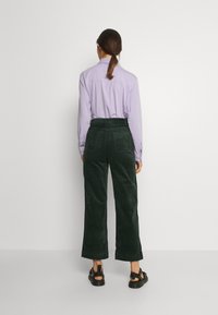 Långärmad, ljuslila skjorta; mörkgröna kordbyxor med vida ben; svarta sandaler; synliga bakfickor och sömmar på byxorna.