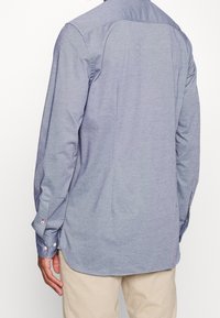 Camisa de manga larga en tejido azul claro con una textura suave, con puños abotonados y dobladillo curvado, mostrada desde atrás.