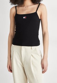 Top aderente nero con spalline sottili e un piccolo logo colorato di Tommy Jeans sul petto. Abbinato a pantaloni a righe color crema.