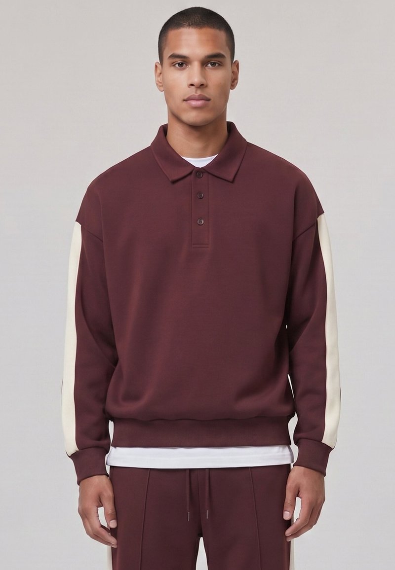 Jeune homme portant un sweat-shirt polo bordeaux à manches longues avec des rayures crème sur les manches et un pantalon bordeaux assorti.