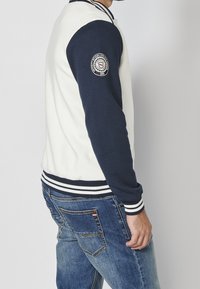 Veste universitaire pour homme en crème et marine. Dotée de poignets et d'un ourlet à rayures côtelées, avec un patch logo sur la manche gauche. Coupe décontractée.