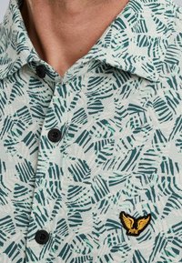 Camisa de manga corta en tejido texturizado color verde menta con un patrón de hojas de palma, que cuenta con un cuello abotonado y un pequeño logo amarillo.