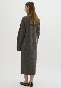 Abito lungo grigio in maglia con colletto alto e spalle scese, che arriva a metà polpaccio. Indossato con scarpe slip-on nere in rete. Design minimalista.
