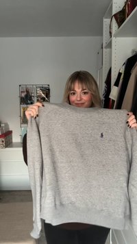 Kobieta o blond włosach trzyma szarą bluzę z małym niebieskim haftowanym logo w pokoju z ubraniami i magazynami.