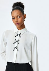 Blusa blanca de manga larga con cuello; presenta tres acentos de cinta negra en la parte delantera. Tejido suave y liso con cierre de botones.