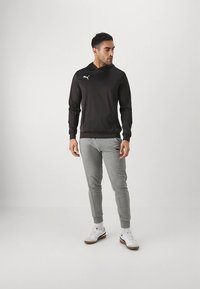 Felpa nera con cappuccio e logo frontale, abbinata a pantaloni da jogging grigio chiaro con fondo stretto e sneakers bianche con dettagli marroni.