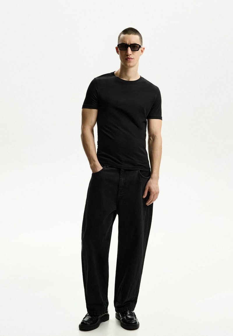 Črna kratka majica s kratkimi rokavi v kombinaciji s širokimi črnimi hlačami in črnimi čevlji. Oblačilo se ponaša z minimalističnim dizajnom in gladko teksturo.