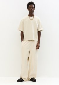 Tricou oversized bej, cu o croială lejeră, asortat cu pantaloni bej cu șnur. Sandale negre cu barete, colier cu lanț completează lookul.