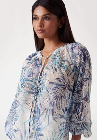 Bluză transparentă cu un model floral în nuanțe de albastru și alb, cu mâneci puff elasticizate, decolteu cu cristal și detalii brodate.