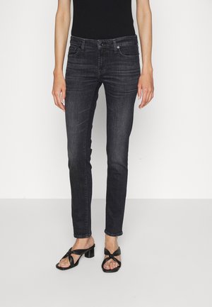 Czarne jeansy slim fit z przednimi kieszeniami, noszone z czarnymi sandałami na obcasie i czarnym, bezrękawnym topem, na jednolitym białym tle.