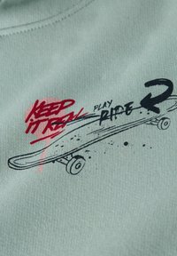 Müntroheline kangas esitleb graafilist disainimustrit, kus on mustad ja punased tekstid "KEEP IT REAL" ning skeitiillustratsioonid plekkide detailidega.