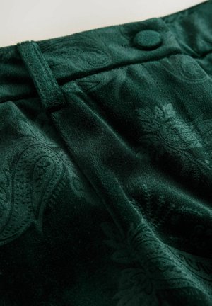Țesătură de catifea verde închis cu modele paisley embosate, un buton acoperit cu țesătură și o buclă pentru brâu pe talia unui articol de îmbrăcăminte.
