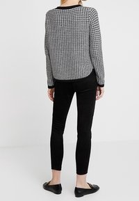 Schwarzer Strickpullover mit strukturiertem Muster, schwarze enganliegende Hose und flache Schuhe. Der Pullover hat lange Ärmel und einen runden Ausschnitt.