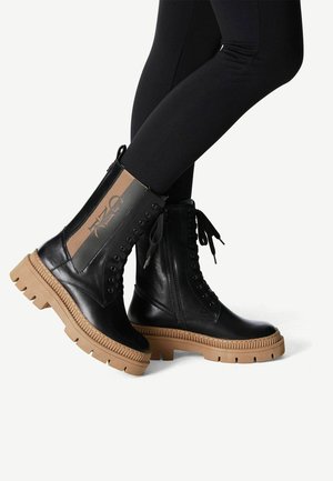 Bottines en cuir noir avec une semelle en caoutchouc beige, à lacets sur le devant et zip sur le côté. Comprend un panneau en tissu marron avec détail de logo.