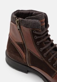 Bottine en cuir marron et daim avec fermeture éclair latérale, col en tissu à motif chevrons et avant rond avec lacets sur semelle noire.