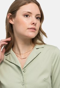 Lichtgroene button-up shirt met een kraag; voorzien van gelaagde roségouden kettingen. Model heeft stijlblond haar en een minimale make-up.