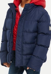 Veste matelassée bleu marine avec une capuche rouge, présentant un design matelassé, une fermeture éclair et des poches latérales, offrant chaleur et confort.
