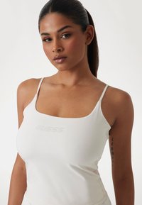 White fitted tank top with thin straps, featuring a subtle logo design on the front. Smooth fabric with a slight sheen.