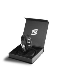 Schwarzes geflochtenes Lederarmband mit silbernen Akzenten in einer schwarzen Geschenkbox. Die Box hat eine glatte, strukturierte Oberfläche und ein kreisförmiges Einlage.