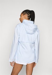 Chaqueta impermeable de color azul claro con capucha, mangas largas y cintura ajustada. Cuenta con una abertura trasera para transpirabilidad y puños elásticos.