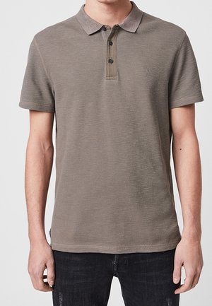 Muž ve hnědém polokošili s krátkými rukávy a třemi knoflíky, v kombinaci s černými džínami, stojí před jednoduchým pozadím.