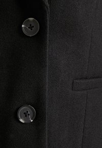 Tessuto di un cappotto di lana nero con due bottoni trasparenti a quattro fori e una tasca a fusello orizzontale sul lato destro.