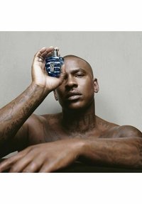 Sinertävä sininen lasinen hajustepullo pidetään paljasta rintaa vasten, jossa näkyy tatuointeja. Pullolla on erottuva muoto ja metallinen suihkeventtiili.