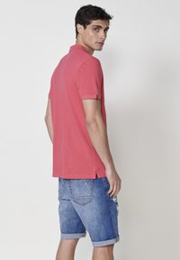 Rdeč bombažni polo majica s kratkimi rokavi, s stranskimi razpoki na robu, v kombinaciji z raztrganimi modrimi denim kratkimi hlačami s zavitimi manšeti.