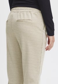 Beige und braune karierte Hose mit elastischem Bund, seitlichen Taschen und glatter Textur, von hinten gezeigt mit einem schwarzen Oberteil.